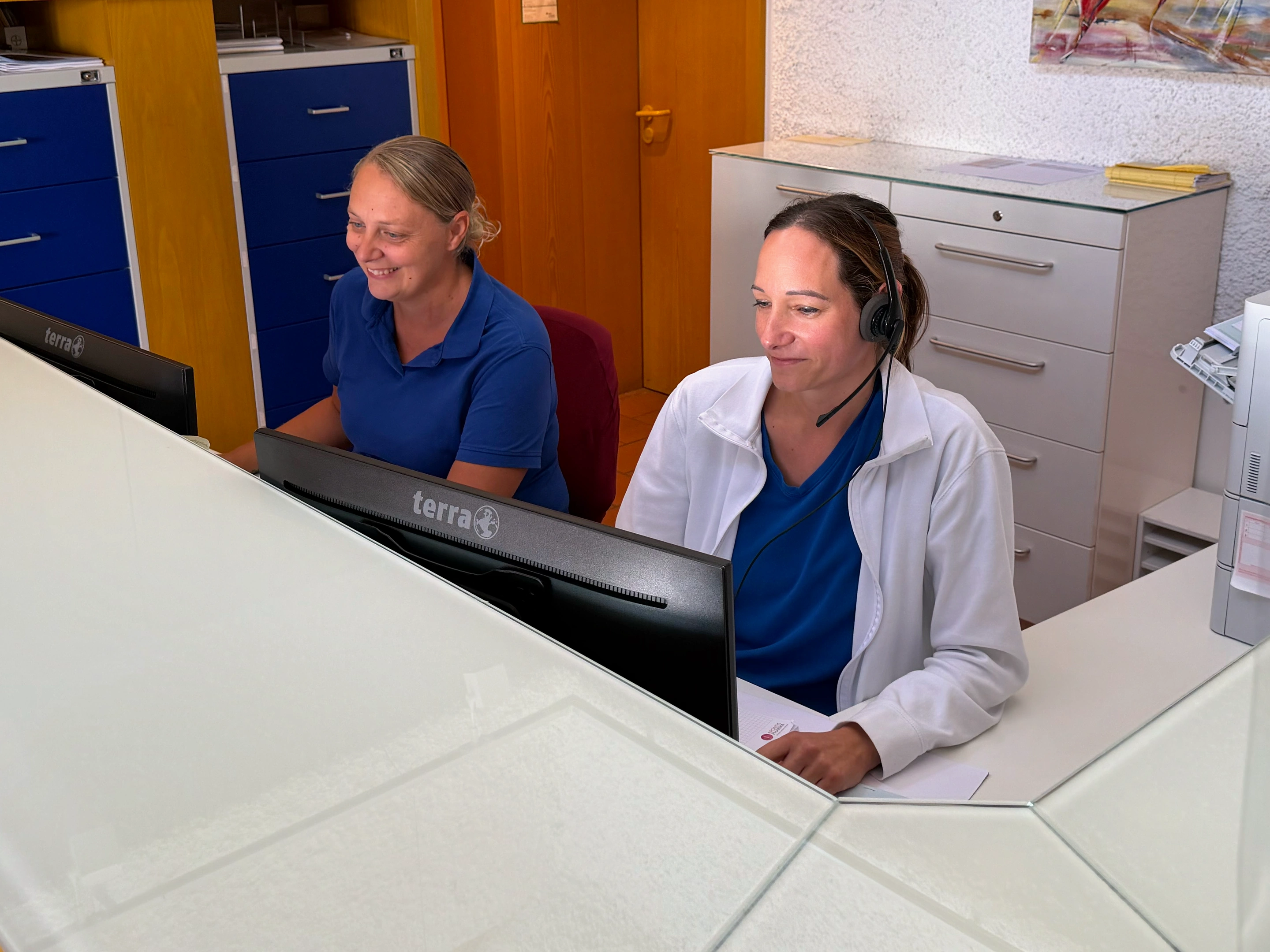 Zwei Mitarbeiterinnen an der Anmeldung, eine davon mit Telefon-Headset.